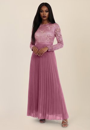 Frau mit langem schwarzen Haar, die ein bodenlanges malvenfarbenes Kleid mit Spitzenoberteil und plissiertem Rock trägt, steht mit vor der Brust verschränkten Händen.