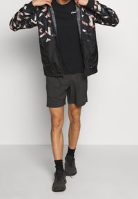 Černá sportovní bunda s barevným geometrickým vzorem, ve spojení s černým tričkem a tmavě šedými šortkami. Model má na sobě černé boty a ponožky.