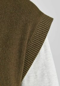 Gros plan d'un pull sans manches en tricot marron superposé sur une chemise à manches longues gris clair sur un fond uni.