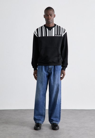 Moletom preto com padrão de riscas verticais brancas, bolso frontal e logótipo "Moschino". Combinado com jeans de ganga azuis de corte largo e sapatos pretos.