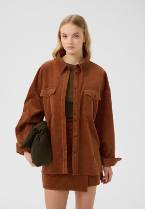 Bruine corduroy overhemd met een kraag, voor knopen en twee borstzakken; gecombineerd met een groene top en een groene tas vastgehouden.