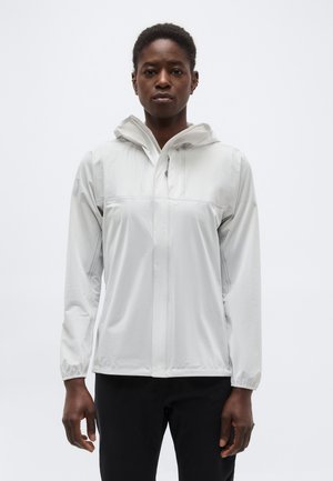 Osoba nosi svetlosivú bundu s kapucňou, predným zipsom, elastickými manžetami a dvoma bočnými vreckami, stojaci pred jednoduchým pozadím.