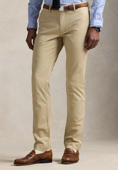 Man die beige broek, bruine riem, bruine leren schoenen, een lichtblauw overhemd en een donkerblauwe stropdas draagt en staat tegen een neutrale achtergrond.