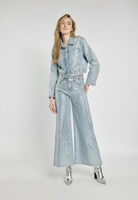 Lśniąca jasnoniebieska krótka kurtka dżinsowa w połączeniu z szerokimi spodniami, obie o metalicznym wykończeniu i subtelnych przetarciach. Srebrne botki za kostkę.