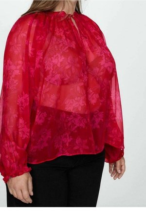 Frau trägt eine transparente rote Bluse mit floralem Muster, langen aufgepufften Ärmeln und schwarze Hose vor einem einfarbigen hellen Hintergrund.