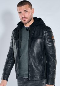 Mann mit kurzem Haar und Bart, trägt eine schwarze Lederjacke über einem dunklen Hoodie und einem grauen Pullover vor hellem, einfarbigem Hintergrund.