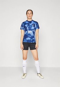Woman standing in blue patterned Tottenham Hotspur soccer jersey, black shorts, white socks, and cream cleats against plain background.
