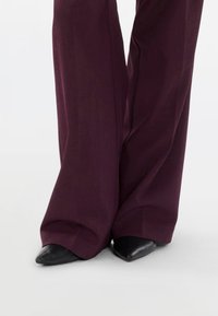 Flared trousers in deep burgundy fabric with a smooth texture, paired with black pointy-toed shoes, showcasing a sleek silhouette.