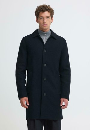 Jeune homme aux cheveux bouclés portant un manteau noir boutonné par-dessus un pull gris à fermeture éclair, debout devant un fond clair uni.