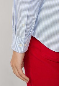 Camisa a rayas azul y blanca con puños abotonados, combinada con pantalones de pana rojos. La camisa presenta rayas finas y una textura suave.
