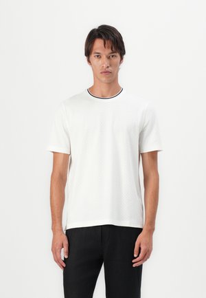 White textured t-shirt with short sleeves, round neck featuring a black and white accent. Paired with dark pants.