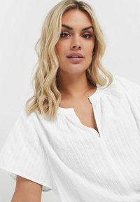 Blusa bianca con scollo rotondo e abbottonatura frontale. Presenta maniche corte e larghe e righe verticali texture. Tessuto leggero.