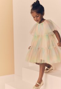 Robe en tulle pastel à design superposé, avec des manches bouffantes courtes et un tissu texturé. Associée à des chaussures dorées brillantes. Se tient sur une plateforme blanche.