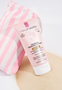 Un tube rose de lotion autobronzante progressive de Brushworks étiqueté "medium", reposant sur du sable à côté d'une pochette en tissu rayée rose et blanche.