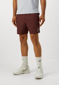 Meeste maroon värvi sportlikud šortid küljetaskutega ja bränditud logosilmaga. Kombineeritud valgete Nike jalanõude ja heledate sokkidega. Sile kangatekstuur.