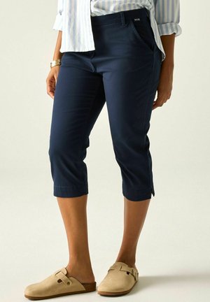 Mujer que lleva pantalones cortos azul oscuro con bolsillos laterales, zapatos beige sin cordones, camisa a rayas y brazalete dorado en la muñeca izquierda.