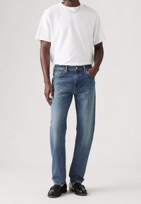 Vaqueros de mezclilla azul con un corte relajado, combinados con una camiseta blanca de manga corta y mocasines negros, de pie contra un fondo neutro.