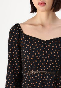 Robe noire à pois orange, décolleté carré, manches bouffantes et dentelle le long du décolleté et de la taille. Tissu doux et texturé.