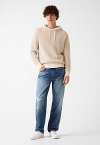Felpa con cappuccio beige in maglia testurizzata con coulisse, abbinata a jeans a gamba dritta blu sbiaditi e sneakers bianche. Stile semplice e contemporaneo.