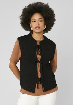 Femme aux cheveux afro naturels portant des boucles d'oreilles blanches, un gilet en tricot noir noué avec trois nœuds sur une chemise marron et un pantalon blanc.