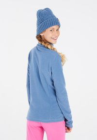 Giacca in pile blu con colletto alto, maniche lunghe e dettagli sulle cuciture posteriori, abbinata a un berretto di lana blu. L'outfit include pantaloni rosa.