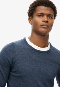 Marineblauwe gebreide trui met een ronde halslijn, voorzien van een witte binnenkraag. De stof heeft een zachte textuur en een subtiel gemêleerd patroon.