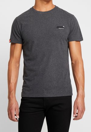 Mann trägt ein eng anliegendes dunkelgraues kurzärmeliges T-Shirt mit kleinem Logo auf der Brust und schwarze Hose vor schlichtem Hintergrund.