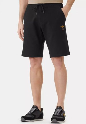 Uomo che indossa pantaloncini neri con coulisse e un piccolo emblema dorato ricamato sulla coscia sinistra, abbinati a scarpe da ginnastica nere con lacci grigi.