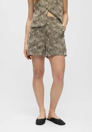 Kobieta ubrana w brązowe szorty z kremowymi, abstrakcyjnymi wzorami spiralnymi i dopasowany top, w połączeniu z czarnymi butami wsuwanymi, stoi z jedną ręką w kieszeni.