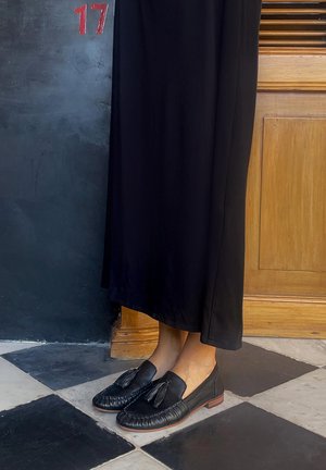 Osoba oblečená v dlhej čiernej sukni a čiernych mokasínach s príveskami stojaca na čierno-bielom štvorcovom podlahe blízko drevených dverí a tmavej steny s číslom 17.