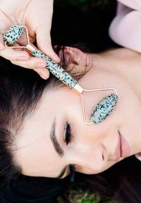 Jade roller do twarzy z nakrapianym zielonym i czarnym kamieniem, złotą metalową rączką, trzymany blisko policzka, oparty na jasnoróżowej powierzchni.