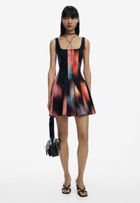 Black sleeveless dress with a fitted bodice, flared skirt, colorful abstract print, and a square neckline; paired with a small black bag.