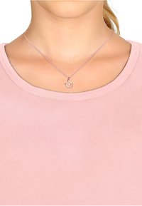 Silvertone halsband med en liten krokhängare, prydd med fem små klara ädelstenar, på en delikat rosa kedja. Bärs över en mjuk rosa skjorta.