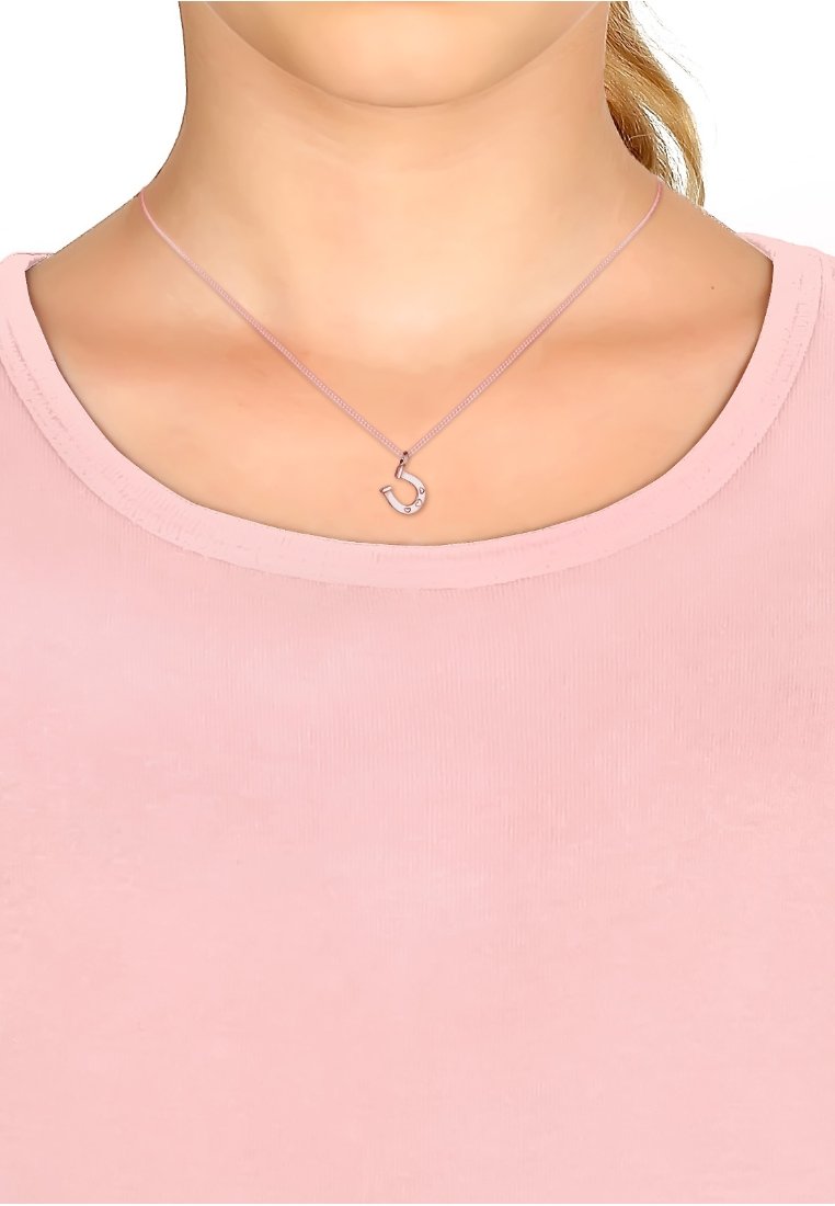 Silvertone halsband med en liten krokhängare, prydd med fem små klara ädelstenar, på en delikat rosa kedja. Bärs över en mjuk rosa skjorta.