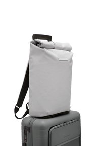 Sac à dos gris à rabat fabriqué en matériau texturé avec des bretelles noires, posé sur une valise grise. Il possède une petite poche avant et une poignée.