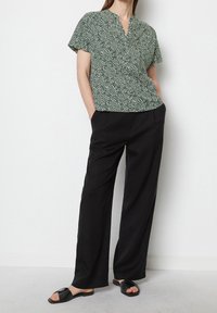 Blusa verde a maniche corte con motivo astratto bianco, abbinata a pantaloni neri a gamba larga e sandali neri scivolati. Tessuto liscio, vestibilità comoda.