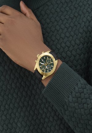 Orologio cronografo dorato con quadrante verde, cinturino in gomma nera e cassa testurizzata. Indossato su un polso sopra a un maglione verde lavorato a maglia.