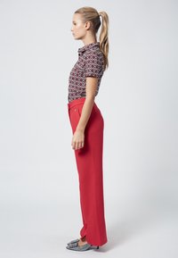 Short-sleeve patterned shirt in black, red, and white, paired with high-waisted red trousers. Footwear features a bow detail and textured accents.