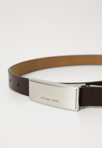 Brauns ādas josts ar spīdīgu sudraba toņa metāla sprādzi, uz kuras ir izreljefēts "MICHAEL KORS." Gluda tekstūra ar precīzi izšūtām šuvēm un caurumiem.