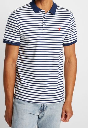 Mies, jolla on yllään laivastonsinivalkoraidallinen lyhythihainen poolopaita, jossa on punainen lintulogo, ja vaaleansiniset farkut, seisoo yksivärisen taustan edessä.