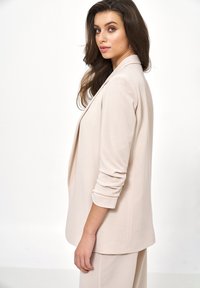 Młoda kobieta z długimi brązowymi włosami, ubrana w beżową marynarkę i pasujące spodnie, stojąca bokiem na białym tle.