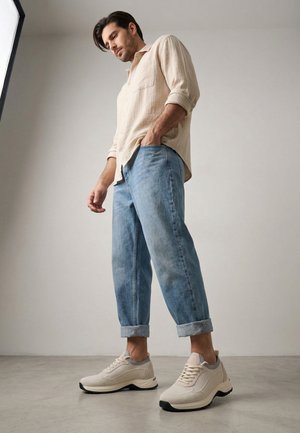 Man draagt een beige overhemd, opgerolde lichtblauwe spijkerbroek en beige sneakers, staand op een grijze vloer tegen een eenvoudige muur.