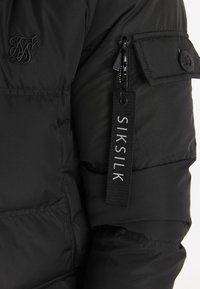 Svart puffjacka med quiltad textur, utrustad med en logotyp på bröstet, en dragkedjeficka och en tagg med "SIKSILK" tryckt i vitt.