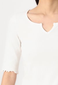 Hvit ribbestrikket topp med V-hals, korte ermer med bølgende kanter, og en tettsittende design. Glatt tekstur uten synlige mønstre.