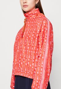 Giacca a vento rossa con motivo, colletto alto, chiusura con zip, polsini elastici e orlo con cordino, caratterizzata da un design riflettente sulle maniche.