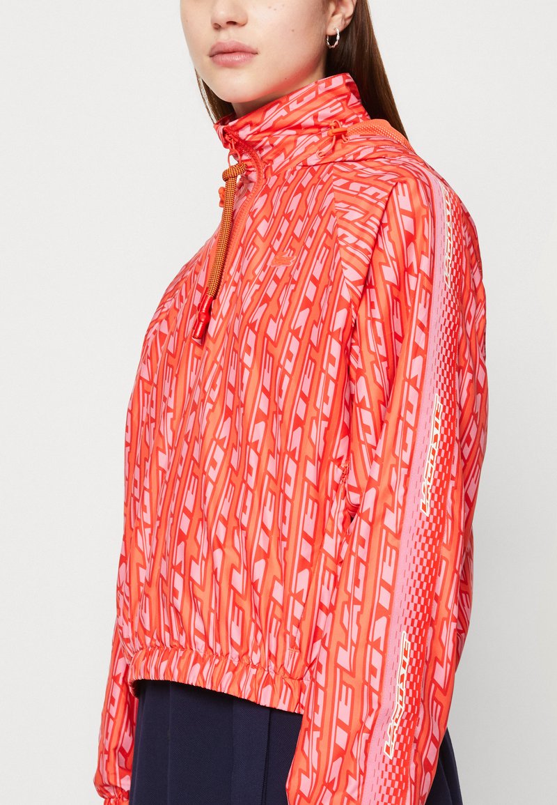 Rompevientos rojo con un patrón, cuello alto, cierre de cremallera, puños elásticos y dobladillo ajustable, que presenta un diseño reflectante en las mangas.