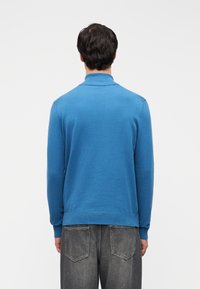 Μπλε πλεκτό πουλόβερ με ριμπ γιακά, μακριά μανίκια και ίσια τελεία, σε συνδυασμό με γκρι τζιν τζιν. Λεπτή υφή, κλασικός σχεδιασμός.