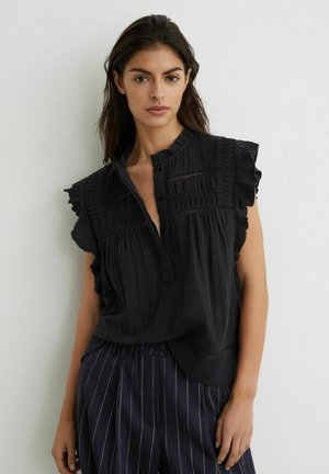 Tânără cu păr lung și închis la culoare, purtând o bluză neagră fără mâneci cu volane și pantaloni bleumarin cu dungi subțiri, stând în fața unui perete simplu.