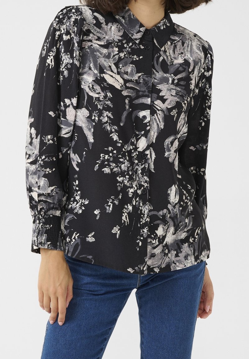 Femme portant un chemisier noir boutonné avec des imprimés floraux blancs et un jean bleu, debout avec une main détendue le long du corps.