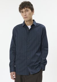 Junger Mann mit zerzaustem braunem Haar, der ein dunkelblaues, gemustertes Hemd über einem weißen Unterhemd trägt und vor einem schlichten weißen Hintergrund steht.